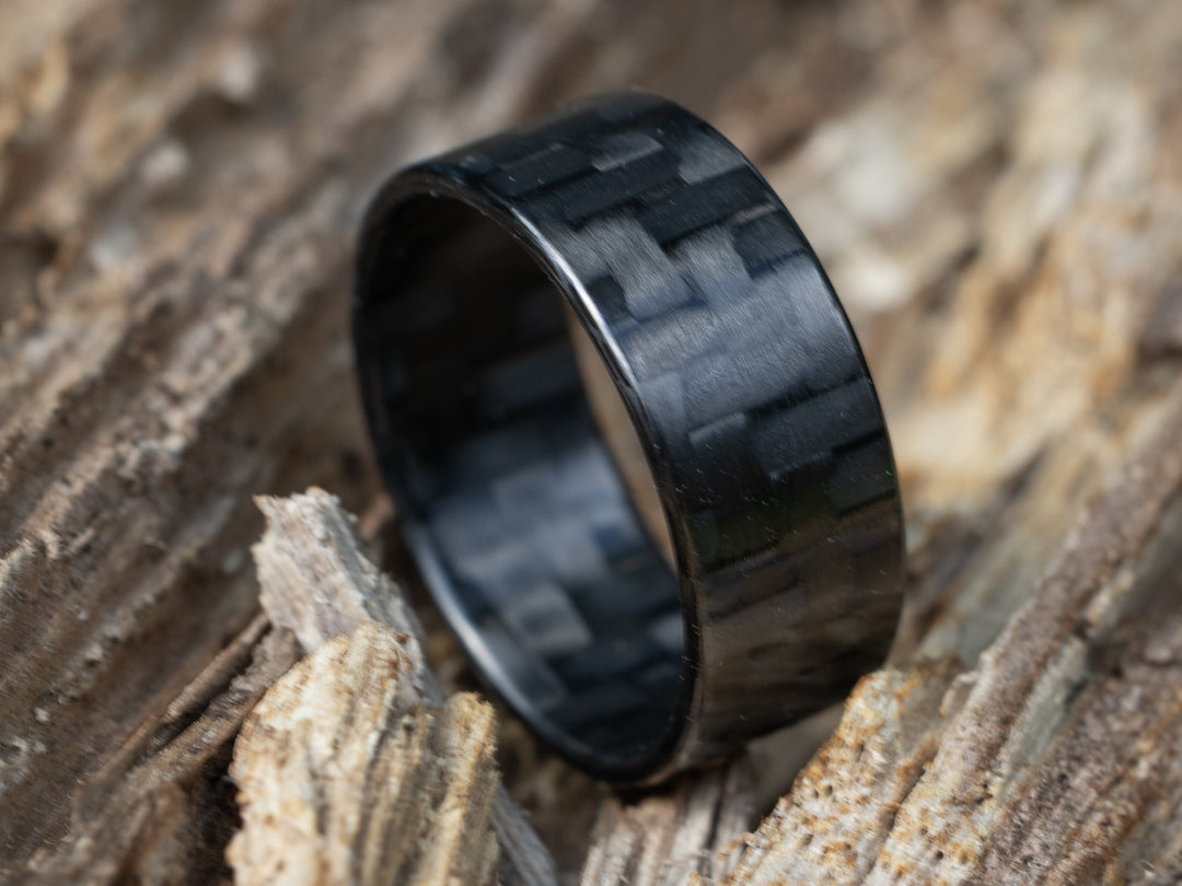 Close-up of 8mm carbon fiber ring with traditional twill pattern, resting on textured tree bark
