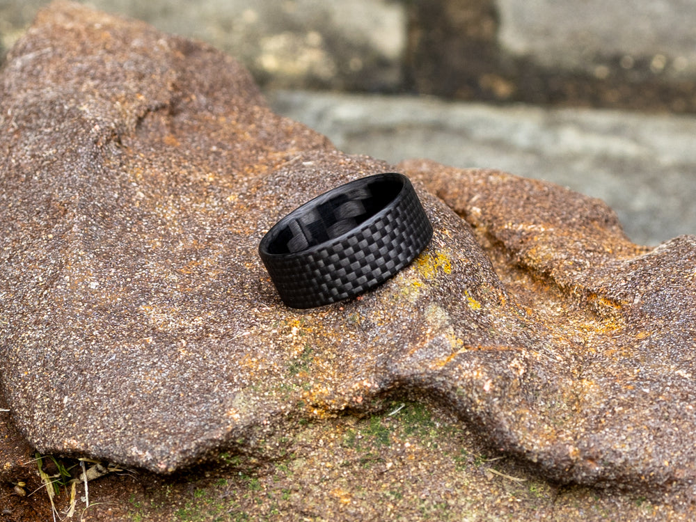 Gridlock 8mm ring resting on a textured rock, highlighting the matte black carbon weave against a rugged outdoor setting