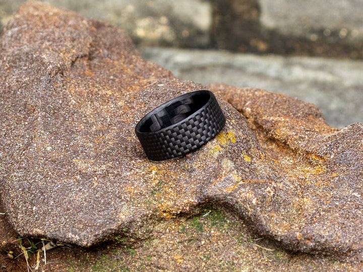 Gridlock 8mm ring resting on a textured rock, highlighting the matte black carbon weave against a rugged outdoor setting