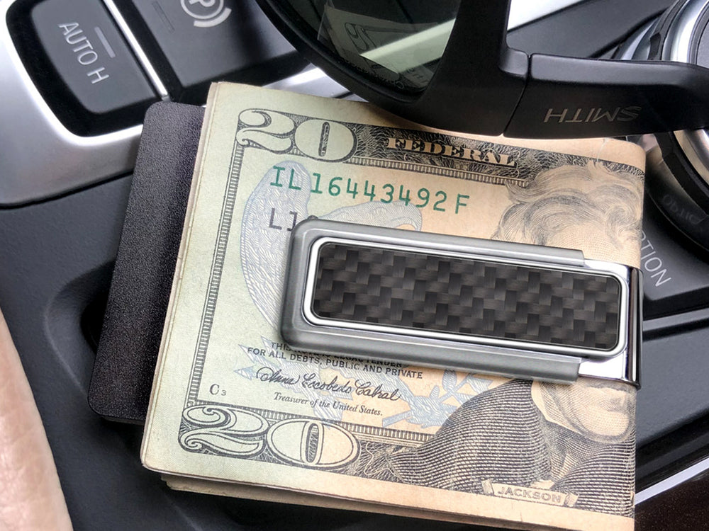 M-Clip stainless steel money clip with black carbon fiber inlay holding cash inside a car interior