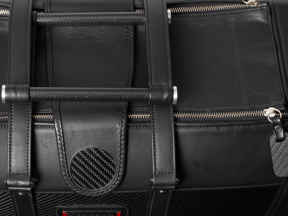 Detailed view of the handles and zipper mechanism on a black leather travel bag with carbon fiber detailing.