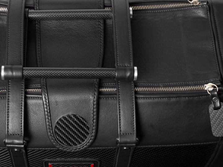 Detailed view of the handles and zipper mechanism on a black leather travel bag with carbon fiber detailing.