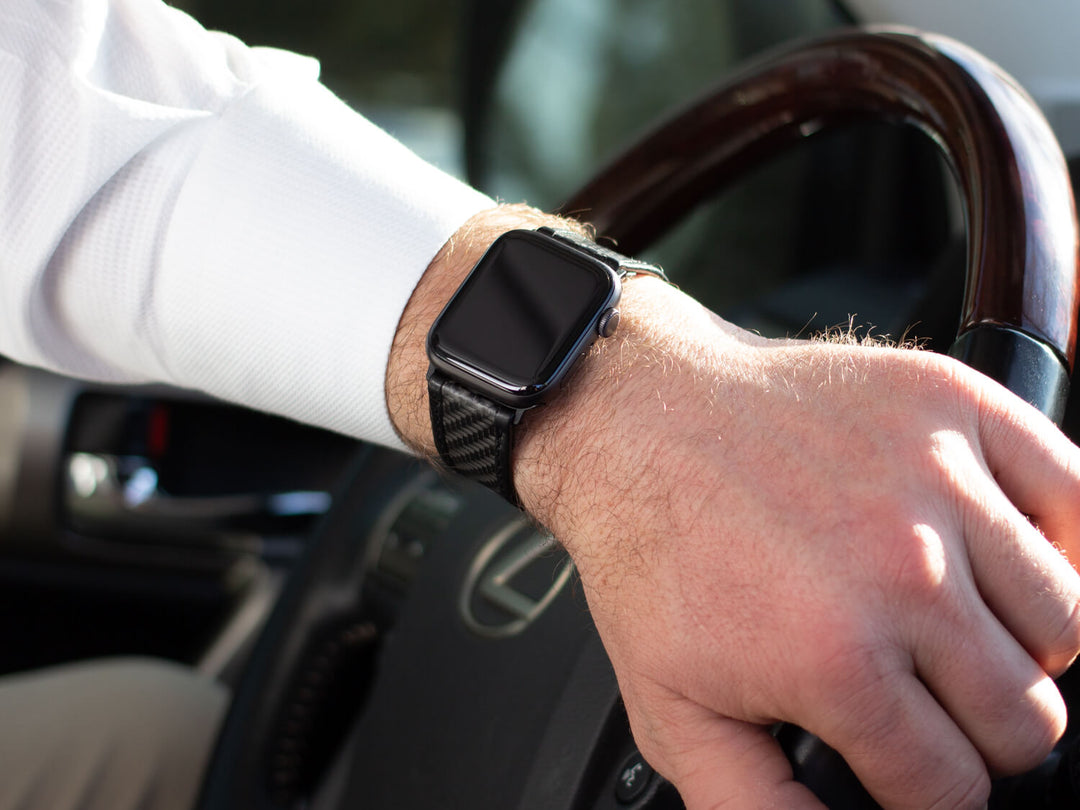 Man's hand on a car steering wheel, wearing an Apple Watch with a CarboBand carbon fiber strap.