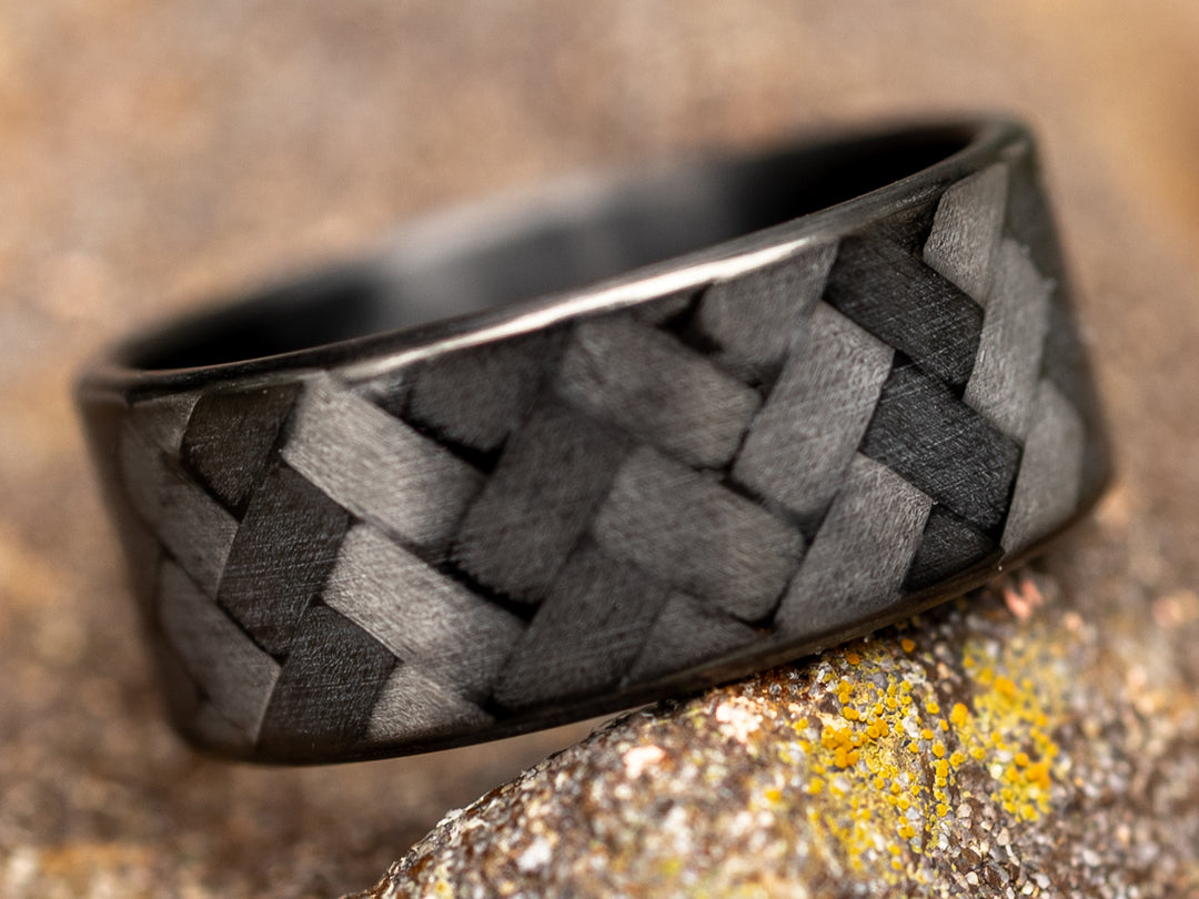8mm black carbon fiber ring with 45-degree weave, photographed on a textured rock surface for a rugged outdoor vibe