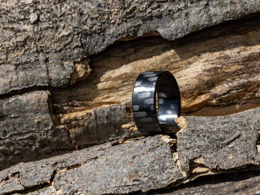 8mm carbon fiber ring with classic twill weave, positioned upright on rough tree bark in bright natural sunlight