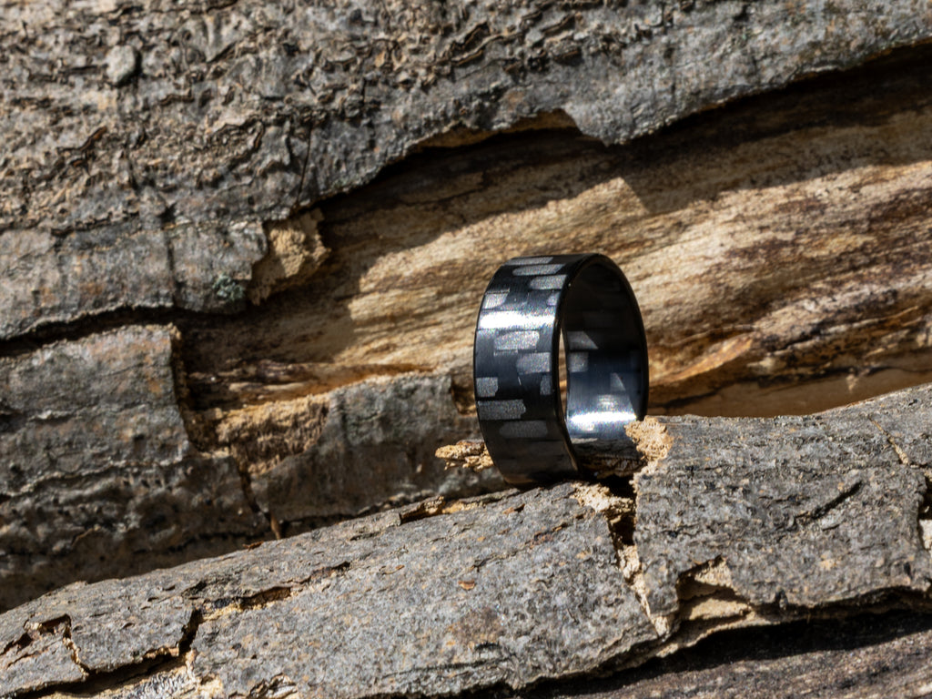 8mm carbon fiber ring with classic twill weave, positioned upright on rough tree bark in bright natural sunlight