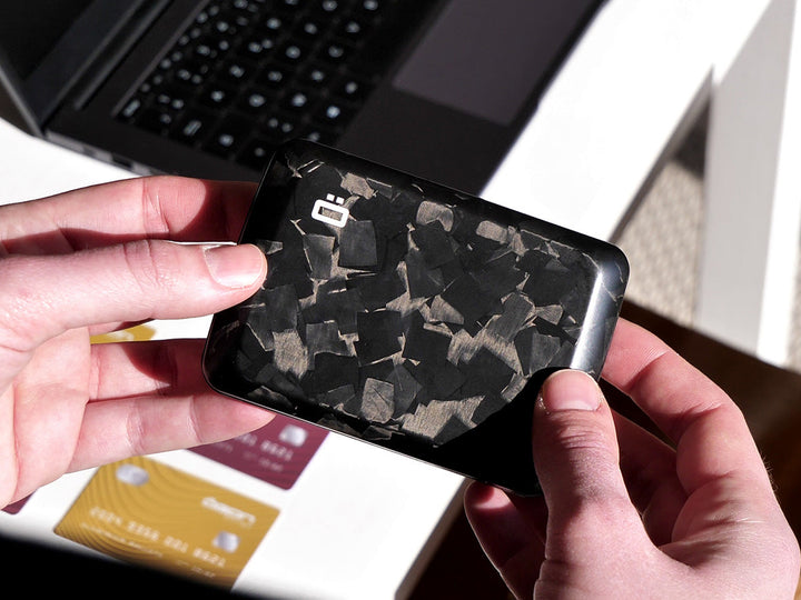 Hands holding a forged carbon fiber wallet with matte marbled texture, shown in natural light near a laptop and credit cards.