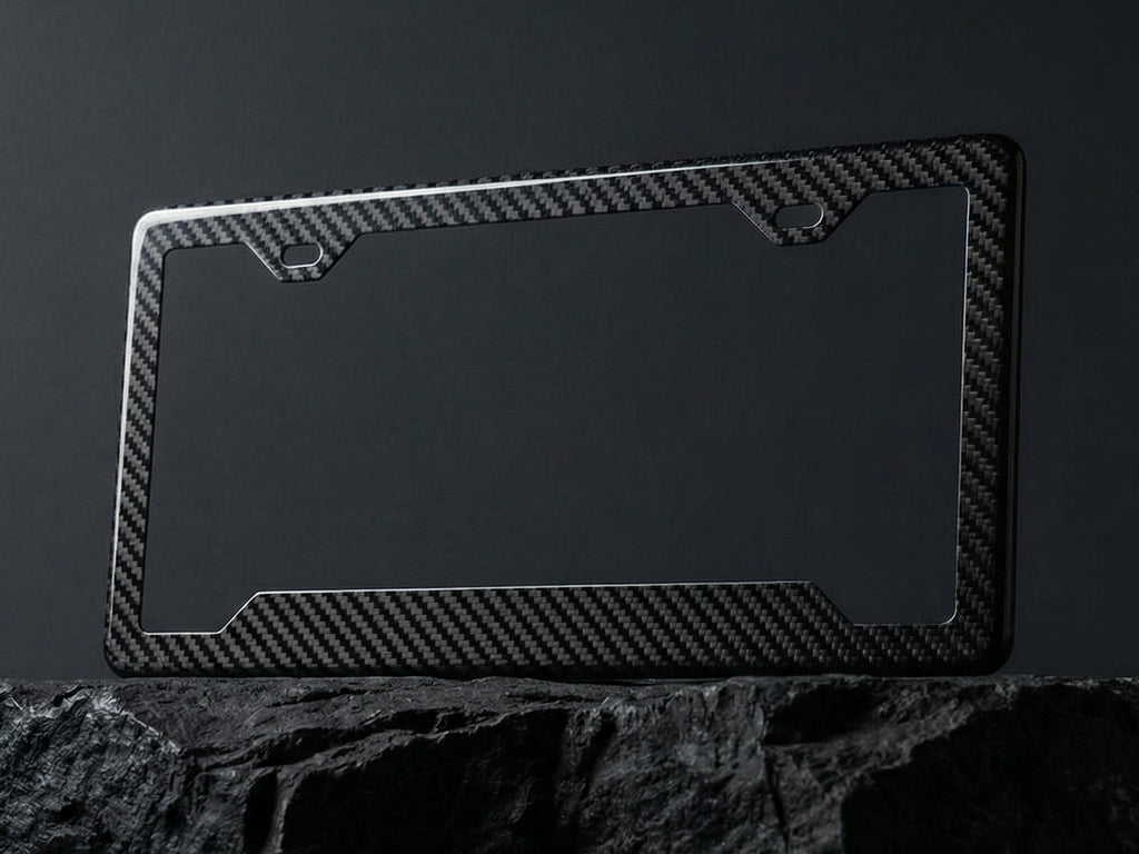 Carbon fiber license plate frame with angled bottom corners displayed upright on a dark stone surface against a charcoal background, showing the twill weave pattern and 2-hole top mounting cutouts