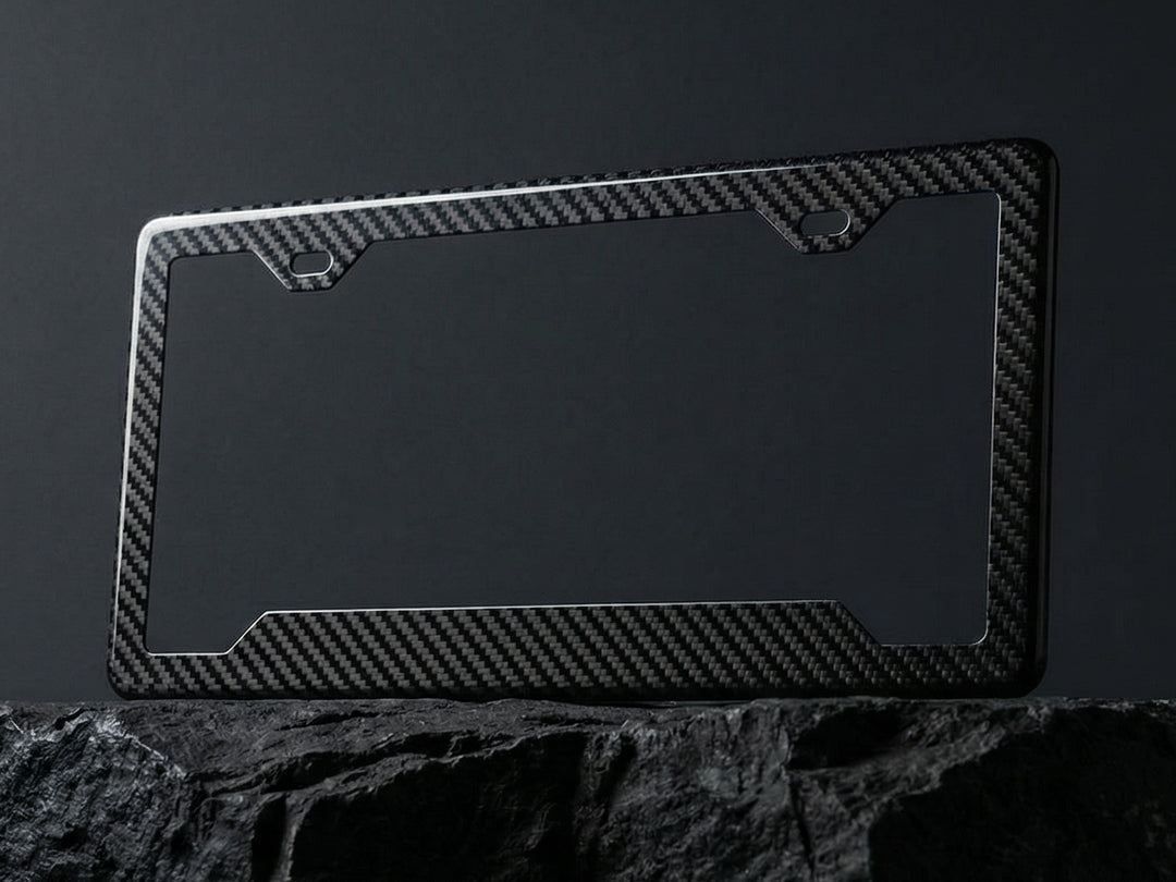 Carbon fiber license plate frame with angled bottom corners displayed upright on a dark stone surface against a charcoal background, showing the twill weave pattern and 2-hole top mounting cutouts