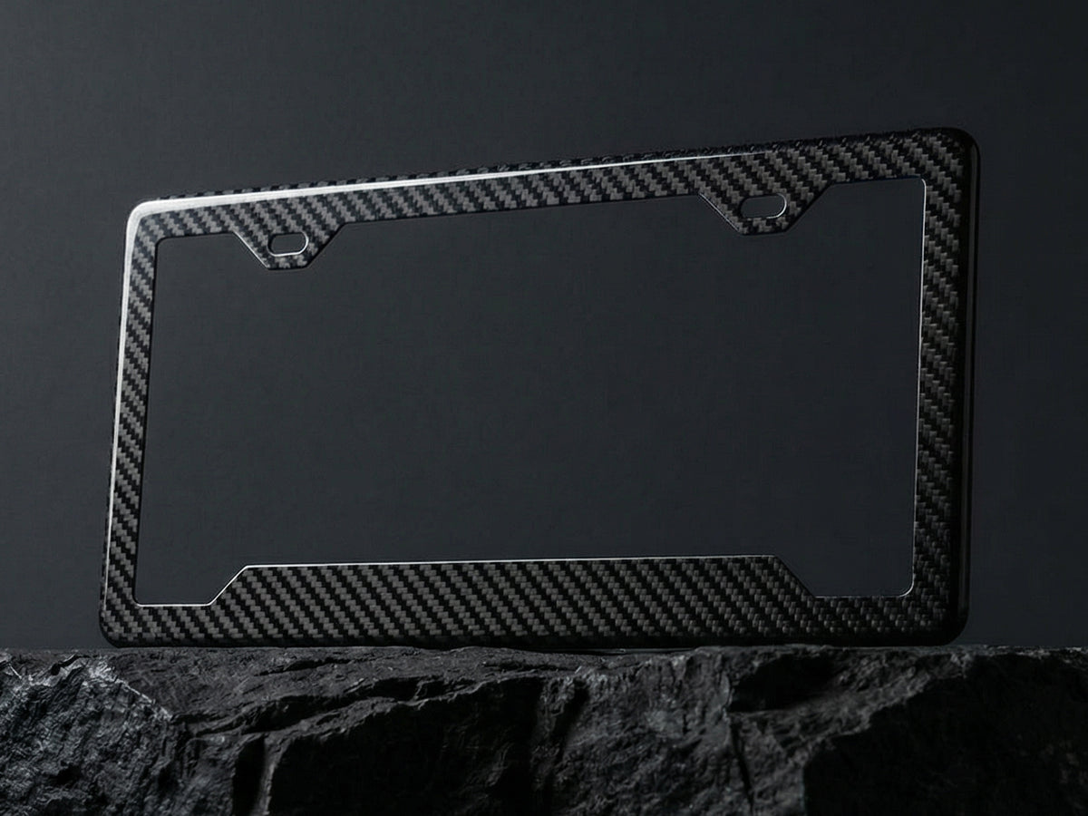 Carbon fiber license plate frame with angled bottom corners displayed upright on a dark stone surface against a charcoal background, showing the twill weave pattern and 2-hole top mounting cutouts