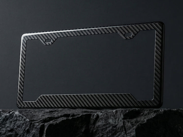 Carbon fiber license plate frame with angled bottom corners displayed upright on a dark stone surface against a charcoal background, showing the twill weave pattern and 2-hole top mounting cutouts