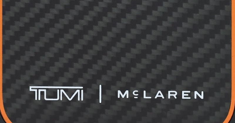 TUMI x McLaren mark in white on the matte carbon fiber back of the iPhone 18 case, with the papaya line at the corner